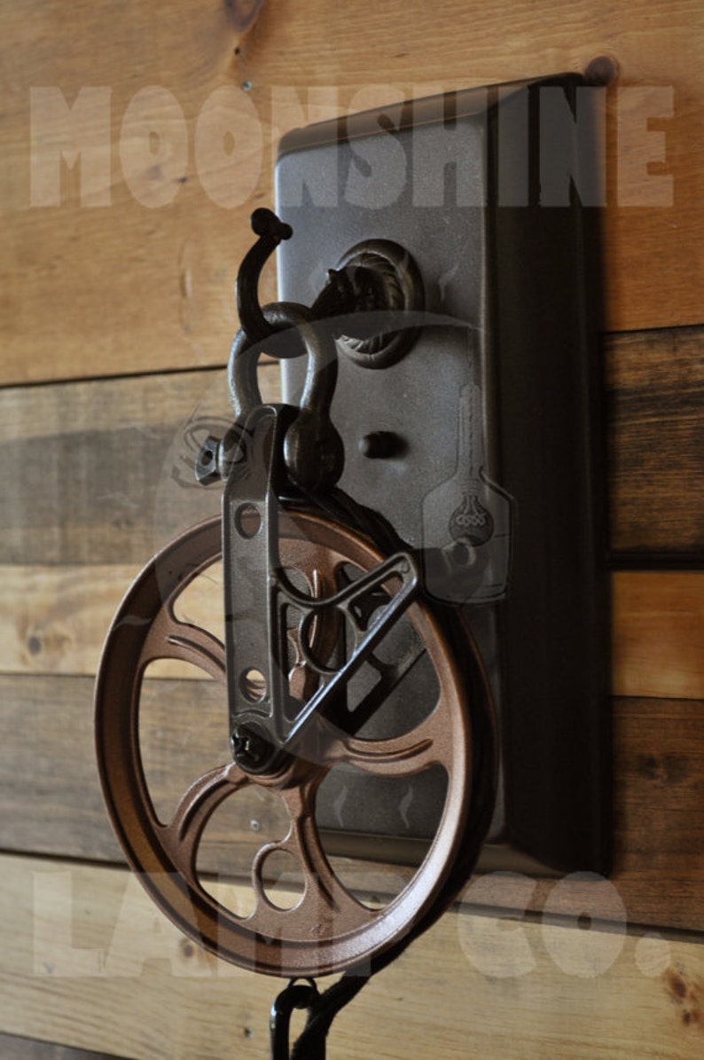 May include: A rustic metal pulley with a brown wooden wheel and a black metal chain. The pulley is mounted on a black metal plate with the text "Moonshine" printed on it.