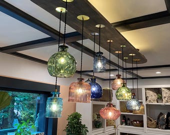 Może przedstawiać: Rustykalna drewniana lampa sufitowa z rzędem kolorowych szklanych lamp wiszących. Lampy są zawieszone na czarnych sznurkach i mają różne kształty i rozmiary. Lampa jest zamontowana do sufitu za pomocą ciemnego drewnianego belki.