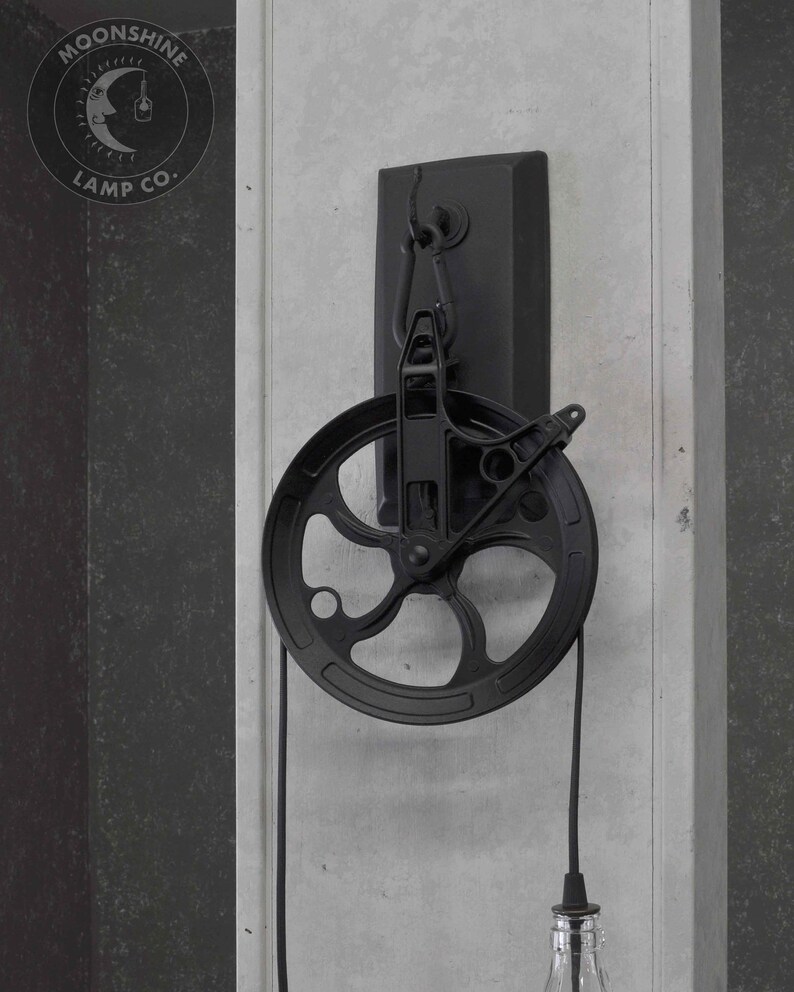 Puede incluir: L&aacute;mpara colgante de estilo industrial negro con una polea y una pantalla de botella de vidrio transparente. La luz est&aacute; montada en una placa de metal negro y tiene un cable negro. La luz est&aacute; etiquetada "Moonshine Lamp Co."
