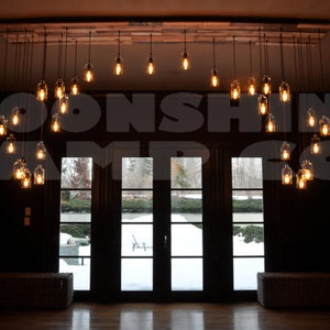 May include: A rustic chandelier made of glass bottles with Edison bulbs hanging from a wooden beam. The chandelier is in a room with a large window overlooking a snowy landscape. The text "Moonshine Lamp Co." is visible in the image.