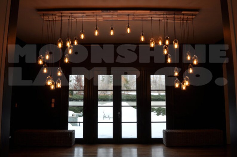 May include: A rustic chandelier made of glass bottles with Edison bulbs hanging from a wooden beam. The chandelier is in a room with a large window overlooking a snowy landscape. The text "Moonshine Lamp Co." is visible in the image.