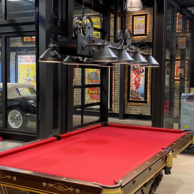 May include: A red felt pool table with a dark wood frame and gold accents. The table is set up in a room with a vintage industrial style, featuring exposed brick walls and a black metal light fixture with multiple shades.