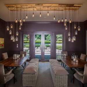 May include: A dining room with a long wooden table and chairs. The room has a dark purple wall and a large window with a view of a garden. A unique chandelier with multiple glass bulbs hangs from the ceiling.