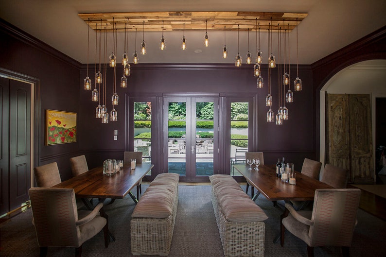 May include: A dining room with a long wooden table and chairs. The room has a dark purple wall and a large window with a view of a garden. A unique chandelier with multiple glass bulbs hangs from the ceiling.
