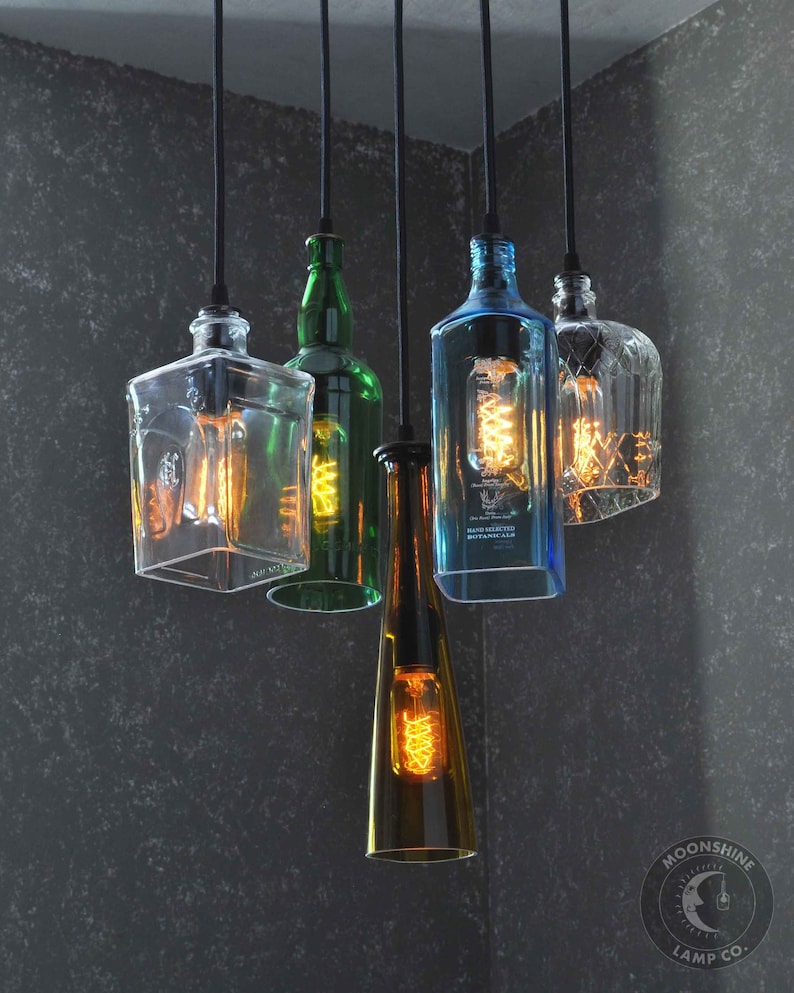May include: A chandelier made from repurposed glass bottles with Edison bulb light fixtures. The bottles are clear, green, blue, and amber. The chandelier is hanging from a black ceiling.