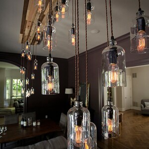 May include: A rustic chandelier with multiple glass bulbs hanging from a wooden beam. The bulbs are lit and cast a warm glow. The chandelier is suspended from the ceiling with brown cords.