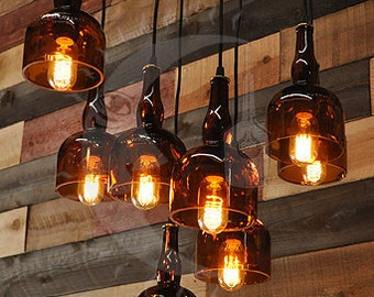 May include: A group of seven amber glass bottle pendant lights hanging from a wooden ceiling. The lights are turned on and illuminate the room with a warm glow.