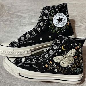 May include: A pair of black high-top canvas trainers featuring white rubber soles and silver eyelets. The side of one shoe displays an embroidered moth, crescent moon, stars, and floral vines, while the other features a circular Converse logo.