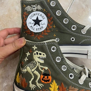 Può includere: Sneakers alte in tela verde oliva con dettagli ricamati. La tomaia presenta un pipistrello bianco, una toppa Converse All Star e uno scheletro di T-Rex che tiene in mano un secchiello a forma di zucca arancione, circondato da foglie autunnali.