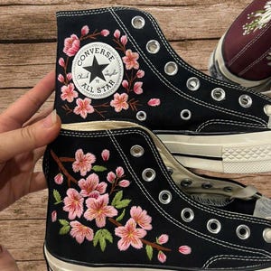 以下が含まれることがあります： ピンクの桜の枝と緑の葉が刺繍された黒いハイカットのキャンバススニーカー。靴の側面には、黒い文字と星のロゴが入った白い円形のコンバースオールスターチャックテイラーのパッチと、銀色のアイレットが付いています。