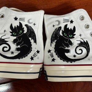 May include: A pair of white high-top canvas trainers featuring a black embroidered dragon illustration with green eyes. The design includes small silver crescent moons and stars. The shoes have silver metal eyelets and a cream rubber sole with red and blue stripes.