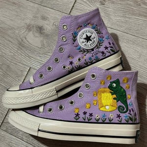 May include: A pair of lavender high-top canvas trainers featuring custom embroidery. One shoe displays a floral wreath surrounding the Converse All Star logo and the text 2026. The other shoe features a green chameleon holding a yellow lantern.