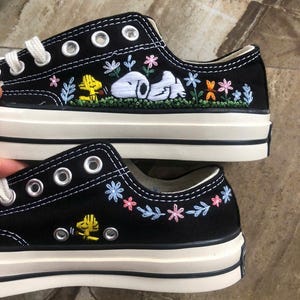 May include: A pair of black low-top canvas trainers featuring white laces and silver eyelets. The side panels are decorated with colourful embroidery of a white dog, a yellow bird, and various small flowers in shades of pink and blue.