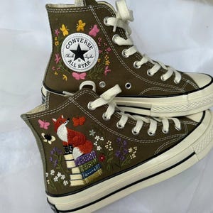 May include: Olive green high-top canvas trainers with white laces and rubber soles. One shoe features a white Converse All Star patch surrounded by embroidered butterflies. The other shoe displays an embroidered fox sitting on a stack of books.