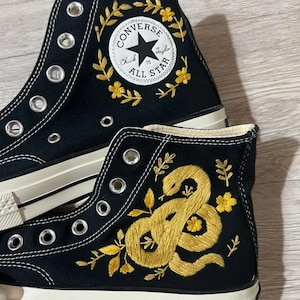 Könnte beinhalten: Schwarze High-Top-Sneaker aus Canvas mit weißen Metallösen und einer cremefarbenen Gummisohle. Das Obermaterial ist mit gelben Blumenkränzen um das Converse All Star-Logo und einer gewundenen Schlange zwischen kleinen gelben Blüten bestickt.