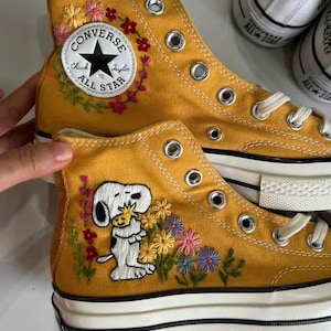 May include: Mustard yellow high-top canvas trainers featuring embroidered floral wreaths around the Converse Chuck Taylor All Star logo patch and a Snoopy character holding Woodstock surrounded by colourful embroidered flowers on the side.