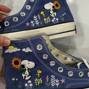 May include: A pair of blue high-top canvas trainers featuring embroidered designs of Snoopy and Woodstock in a garden setting. The embroidery includes a yellow sunflower, various small flowers, a sun, and clouds. The shoes have white rubber soles.