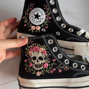 May include: A pair of black high-top canvas sneakers featuring white laces and silver metal eyelets. The upper shoe displays the circular white Converse All Star logo patch. The lower shoe features an embroidered skull surrounded by pink flowers.