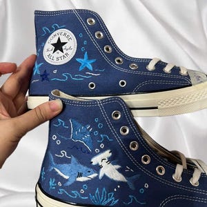 Puede incluir: Un par de zapatillas altas de lona azul marino con bordados personalizados de vida marina, incluyendo un tiburón, un tiburón martillo y una raya. Tienen cordones blancos, ojales de metal plateado y una suela de goma blanca con borde negro.