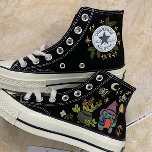 Peut inclure: Une paire de baskets montantes en toile noire avec des lacets blancs et des semelles en caoutchouc. Les chaussures présentent des broderies personnalisées incluant une grenouille verte avec un chapeau violet, un chaudron, des plantes, des étoiles et un croissant de lune. Le logo Converse All Star est visible.