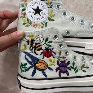 Könnte beinhalten: Ein hellgrüner High-Top-Sneaker aus Canvas mit gestickten Insekten, darunter eine Biene, ein roter Marienkäfer, ein blau-gelber Käfer und ein violetter Käfer. Der Schuh zeigt einen weißen Converse Chuck Taylor All Star Aufnäher mit Blumenstickerei.