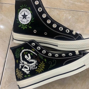 Könnte beinhalten: Ein Paar schwarze High-Top-Sneaker aus Canvas mit weißer Gummisohle und silbernen Ösen. Ein Schuh zeigt das weiße, runde Converse All Star-Logo mit grüner Stickerei. Der andere Schuh ist mit einem weißen, gestickten Dinosaurierskelett verziert.