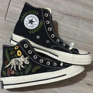 Puede incluir: Un par de zapatillas altas de lona negra con suelas y punteras de goma blanca. Una zapatilla presenta un parche circular blanco de Converse All Star con bordado de hojas verdes. La otra muestra un esqueleto de dinosaurio blanco bordado.