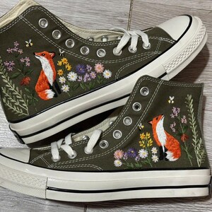 May include: A pair of olive green high-top canvas sneakers featuring detailed embroidery of a red fox sitting among colorful wildflowers and small bees. The shoes have white laces, silver metal eyelets, and a white rubber sole with a black stripe.