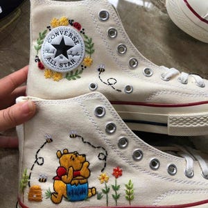 Puede incluir: Zapatillas altas de lona color blanco roto con detalles bordados. El zapato superior muestra un parche circular Converse All Star rodeado de flores amarillas y mariquitas. El zapato inferior presenta un personaje de Winnie the Pooh bordado con abejas, una colmena y flores coloridas.