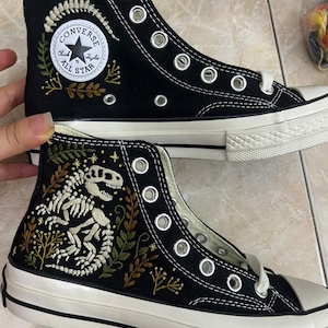 Op de afbeelding: Een paar zwarte hoge canvas sneakers met witte borduursels van een dinosaurusskelet en botanische elementen. De schoenen hebben witte rubberen zolen, zilverkleurige metalen oogjes en een ronde witte Converse All Star patch op de zijkant.