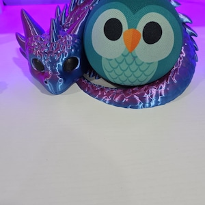 May include: A 3D printed articulated dragon with a metallic blue and purple gradient finish, featuring large dark eyes and textured scales, coiled around a spherical smart speaker designed with a teal owl face, large white eyes, and an orange beak.