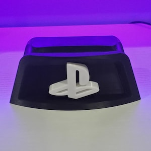 Op de afbeelding: Een zwarte plastic controllerstandaard met een wit PlayStation-logo in het midden van de voorzijde. De standaard heeft een brede, gebogen voet en een uitsparing aan de bovenzijde om een gamecontroller stevig op zijn plaats te houden.