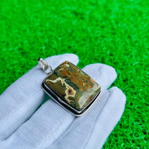 May include: A rectangular pendant featuring a polished, multi-colored stone with earthy tones of green, brown, and cream, set in a smooth silver-toned metal frame with a small circular bail at the top for attachment to a chain or cord.