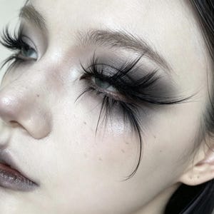 Puede incluir: Vista de cerca de pestañas postizas negras, largas y finas aplicadas en los párpados superiores. El maquillaje presenta un difuminado de sombra de ojos gris ahumado en los párpados, combinado con un labial oscuro y mate en los labios.