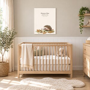 Storybook hedgehog canvas in a nursery on a greige wall above a wooden crib and white wainscoting, showing sample name Eleanor Mae and birth details.