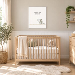 Baby elephant canvas in a nursery on a greige wall above a wooden crib and white wainscoting, showing sample name Eleanor Mae and birth details.