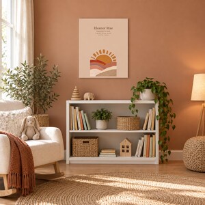 Boho sun canvas in a nursery on a terracotta wall above a white bookshelf, showing sample name Eleanor Mae and birth details.