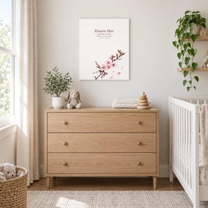 Cherry blossom canvas in a nursery above a light oak dresser beside a white crib, showing sample name Eleanor Mae and birth details.