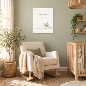 Baby elephant canvas in a nursery on a sage green wall above a cream rocking chair, showing sample name Eleanor Mae and birth details.