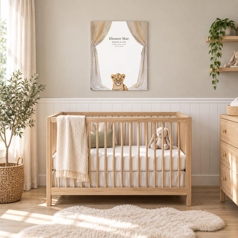 Lion cub canvas in a nursery on a greige wall above a wooden crib and white wainscoting, showing sample name Eleanor Mae and birth details.