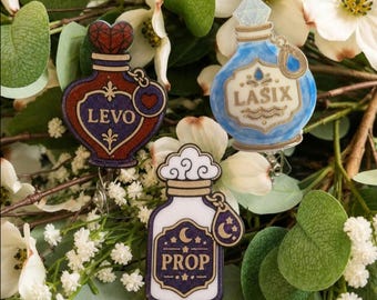 May include: Three decorative badge reels shaped like potion bottles. One is dark red with a heart topper and the text LEVO. One is light blue with a diamond-shaped topper and the text LASIX. One is white and purple with the text PROP.