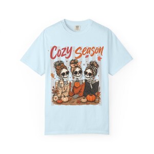 May include: A light blue t-shirt featuring a graphic of three skeletons with messy buns and sunglasses, wearing sweaters and holding drinks. The text Cozy Season is printed above them in orange. The design includes pumpkins and autumn leaves.