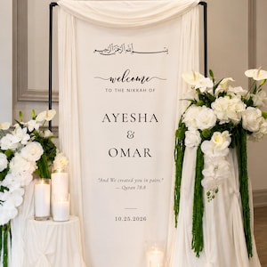 May include: A white wedding welcome sign on a black metal frame, draped with white fabric. The sign features black text: "welcome TO THE NIKKAH OF AYESHA & OMAR", "And We created you in pairs." - Quran 78:8, and "10.25.2026". White floral arrangements and pillar candles surround the base.