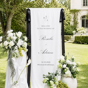 May include: A vertical white fabric wedding welcome sign featuring black text that reads Welcome to the wedding of Rosalie and Adrian, the date 10.10.2026, and the hashtag #ForeverAndAlways. The banner is accented with black ribbons.