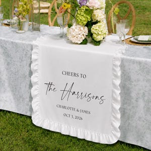 Peut inclure: Un chemin de table blanc à volants avec le texte imprimé CHEERS TO The Harrisons CHARLOTTE & JAMES OCT 3, 2026. Le chemin est posé sur une longue table avec une nappe grise à motifs, entourée de chaises en bois et de centres de table floraux.