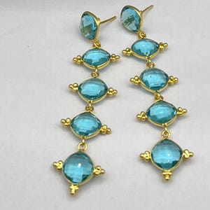 Peut inclure: Une paire de longues boucles d'oreilles pendantes composées de quatre pierres bleu clair à facettes de forme carrée, serties dans un métal doré. Chaque pierre est ornée de petits détails décoratifs dorés aux coins, formant un design vertical.