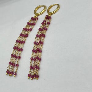 Puede incluir: Un par de pendientes de aro dorados con tres cadenas colgantes en cada uno. Cada cadena está adornada con pequeñas cuentas facetadas de color rosa intenso. Los pendientes tienen un cierre de bisagra y un diseño delicado y superpuesto.