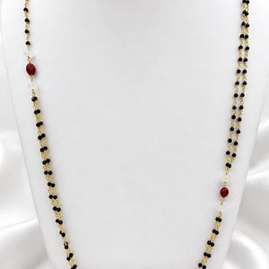May include: A long double-strand necklace featuring small black beads and gold-toned chain links. The design is accented with intermittent clear white beads and oval-shaped deep red beads spaced evenly along the length of the two delicate chains.