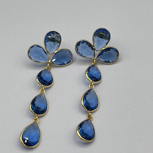 Peut inclure: Une paire de boucles d'oreilles pendantes dorées composées de trois cristaux bleus facettés en forme de poire disposés en trèfle, suivis d'une chaîne verticale de trois autres cristaux bleus en forme de poire suspendus.