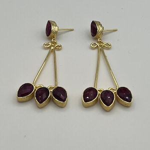 Può includere: Un paio di orecchini pendenti color oro con una singola pietra rotonda color rubino sul perno e tre pietre a forma di pera color rubino sospese da una struttura a filo triangolare con un occhiello decorativo sulla parte superiore.